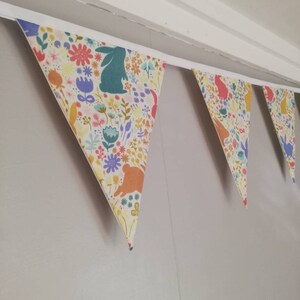 May include: A bunting with a white background and a colorful floral pattern featuring bunnies. The bunting is made of fabric and has a white string for hanging.