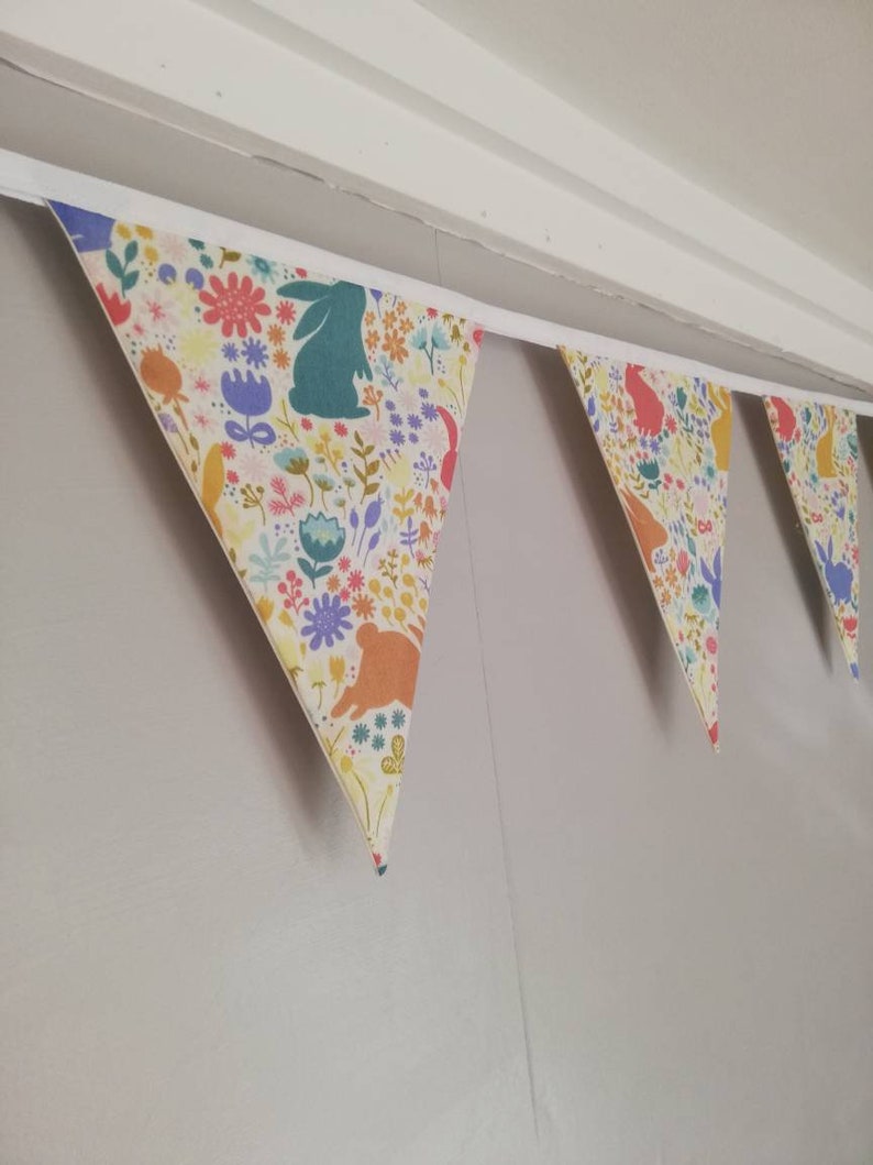 May include: A bunting with a white background and a colorful floral pattern featuring bunnies. The bunting is made of fabric and has a white string for hanging.