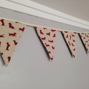 May include: A bunting with a pattern of red dachshund silhouettes on a beige background. The bunting is made of fabric and has a white ribbon running through it.