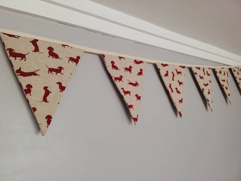 May include: A bunting with a pattern of red dachshund silhouettes on a beige background. The bunting is made of fabric and has a white ribbon running through it.