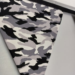 Puede incluir: Una bandera de tejido de camuflaje gris, negro y blanco. La bandera tiene forma triangular y est&aacute; colgada de una cuerda negra.