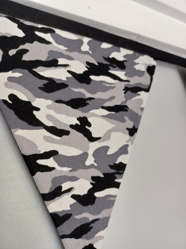 Puede incluir: Una bandera de tejido de camuflaje gris, negro y blanco. La bandera tiene forma triangular y est&aacute; colgada de una cuerda negra.
