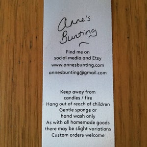 May include: White tag with black text. The text reads "Anne's Bunting" followed by contact information: "Find me on social media and Etsy www.annesbunting.com annesbunting@gmail.com". The tag also includes care instructions: "Keep away from candles / fire Hang out of reach of children Gentle sponge or hand wash only As with all homemade goods there may be slight variations Custom orders welcome"