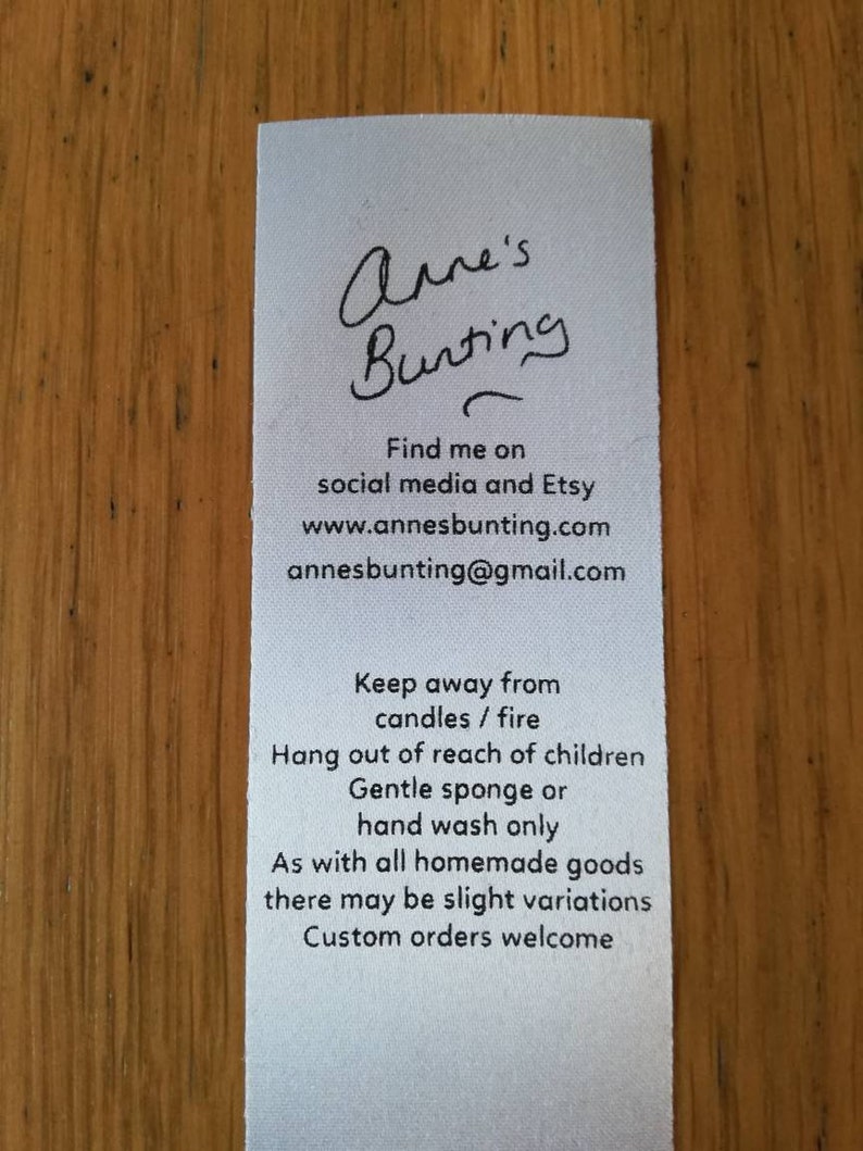 May include: White tag with black text. The text reads "Anne's Bunting" followed by contact information: "Find me on social media and Etsy www.annesbunting.com annesbunting@gmail.com". The tag also includes care instructions: "Keep away from candles / fire Hang out of reach of children Gentle sponge or hand wash only As with all homemade goods there may be slight variations Custom orders welcome"