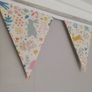 May include: A bunting with a white background and a colorful floral pattern. The bunting has a blue bunny, a yellow bunny, and pink and yellow flowers. The bunting is hanging from a white ceiling.