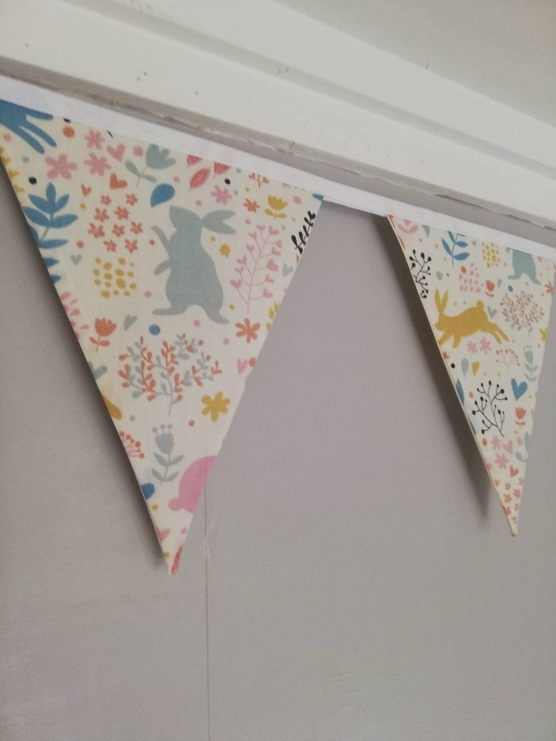 May include: A bunting with a white background and a colorful floral pattern. The bunting has a blue bunny, a yellow bunny, and pink and yellow flowers. The bunting is hanging from a white ceiling.