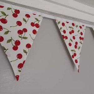 May include: A white bunting with red cherries printed on it. The bunting is strung up on a white ceiling.