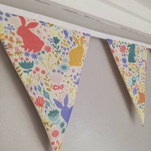 May include: A white fabric bunting with a floral pattern and bunny silhouettes in various colors, including pink, yellow, blue, and green. The bunting is hung on a white ceiling.