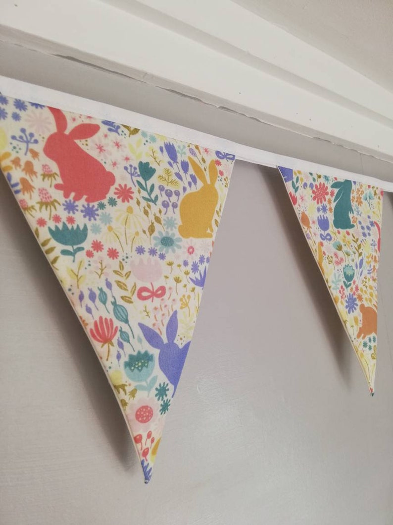 May include: A white fabric bunting with a floral pattern and bunny silhouettes in various colors, including pink, yellow, blue, and green. The bunting is hung on a white ceiling.