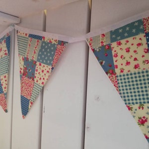 May include: A patchwork fabric bunting with alternating blue, pink, and cream triangles. Each pennant features a mix of floral, polka dot, and gingham patterns. The bunting is strung on a white ribbon, perfect for adding a touch of vintage charm to any space.