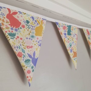May include: A colorful fabric bunting with a white background and a repeating pattern of bunnies and flowers. The bunting is made of triangular flags and is strung together with white ribbon.