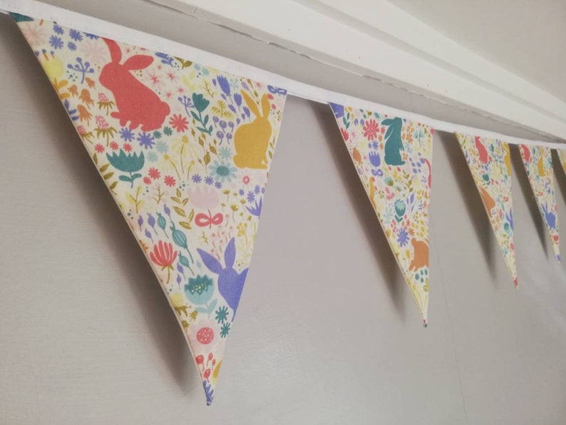 May include: A colorful fabric bunting with a white background and a repeating pattern of bunnies and flowers. The bunting is made of triangular flags and is strung together with white ribbon.
