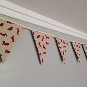 May include: A beige fabric bunting with red dachshund silhouettes. The bunting is strung up against a white ceiling.