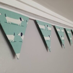 May include: A green and white bunting with a repeating pattern of dachshunds. The bunting is strung up on a white string and is hanging from a white ceiling.