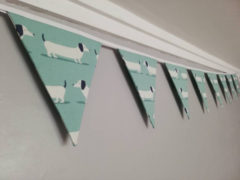 May include: A green and white bunting with a repeating pattern of dachshunds. The bunting is strung up on a white string and is hanging from a white ceiling.