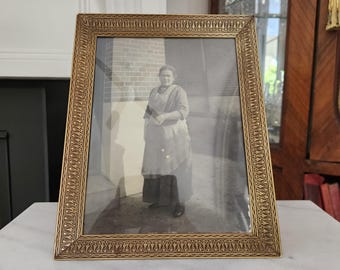 May include: A framed black and white photograph featuring a woman in a long dress and apron. The frame is gold-colored with an ornate, repeating pattern. The photograph is displayed on a white marble surface.