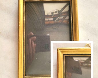 May include: A rectangular, gold-colored picture frame with a detailed border and a small hanging loop. The frame surrounds a reflective surface, with a smaller inset frame in the lower right corner. The frame is displayed against a light-colored background.