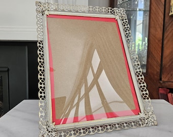 May include: A vintage silver-toned picture frame with a decorative, openwork border. The frame has a red mat and a clear glass cover. The frame is angled, showing the reflection of a window. A classic piece for displaying photos or artwork.