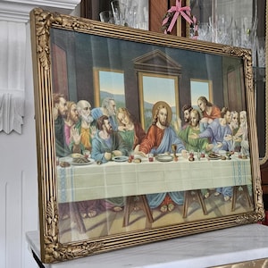 May include: A framed print of The Last Supper, depicting Jesus and his disciples. The artwork is in a gold-coloured ornate frame. The figures are in robes, and the table is set with food and drink. The background is a dark blue.