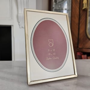 May include: A rectangular gold-toned picture frame with an oval-shaped maroon background. The frame has the text "9 x 12" and "10 x 15" and "Tyden Quality". The frame is displayed on a marble surface.