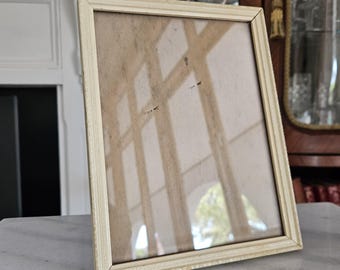 May include: A rectangular, off-white picture frame with a glass front. The frame is made of wood and has a decorative, ridged border. The glass reflects the surrounding environment. The frame is displayed on a marble surface.