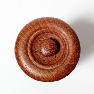 May include: A close-up of a wooden pepper mill top with a circular design and a small knob in the center. The top has a dark brown color and a smooth, polished finish.