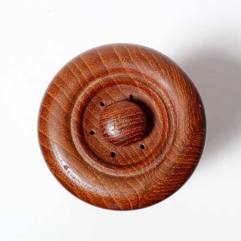 May include: A close-up of a wooden pepper mill top with a circular design and a small knob in the center. The top has a dark brown color and a smooth, polished finish.