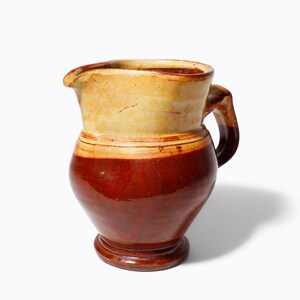 May include: A ceramic jug with a handle, featuring a two-tone design. The top section is a light beige, transitioning to a deep reddish-brown on the lower half. The jug has a spout for pouring.