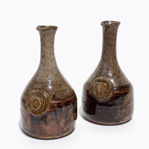 May include: Two handcrafted ceramic bottles with a rustic aesthetic. The bottles feature a brown and tan colour palette with a textured surface. Each bottle has a circular emblem with a letter. The bottles are approximately 20 cm tall.