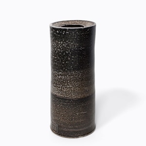May include: A tall, cylindrical vase with a dark, speckled exterior. The vase has a slightly textured surface and a dark brown base. The top opening is a dark, circular shape. The vase is a decorative item.