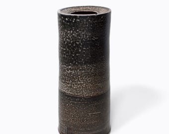 Könnte beinhalten: Eine hohe, zylindrische Vase mit dunkler, gesprenkelter Außenseite. Die Vase hat eine leicht strukturierte Oberfläche und einen dunkelbraunen Boden. Die obere Öffnung ist dunkel und kreisförmig. Die Vase ist ein dekorativer Gegenstand.