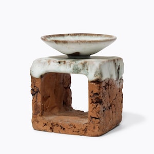 Peut inclure: Sculpture en céramique faite à la main, composée d'un petit bol peu profond posé sur une base carrée ajourée. La base est brun-rouge avec une surface texturée, tandis que le dessus et le bol sont émaillés blanc et vert clair.