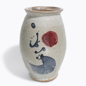 May include: A speckled, off-white ceramic vase with a wide mouth and rounded body. The vase is decorated with a red circle, dark blue brushstrokes, and a dark blue wave design. The vase has a rustic, handmade appearance.