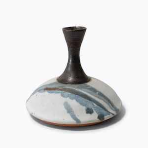 May include: A handcrafted ceramic vase with a broad, flat base and a tall, slender neck. The base is white with blue and brown streaks. The neck is dark brown. The vase is a unique piece of art.