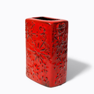 May include: A rectangular, red ceramic vase with a glossy finish. The vase has a repeating pattern of stylized floral designs carved into its surface. The interior of the vase is black. A decorative item.
