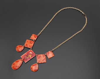 May include: A gold-toned chain necklace with a pendant featuring geometric shapes in shades of orange and red. The pendant has a central square with a patterned design, and other shapes include squares and teardrops.