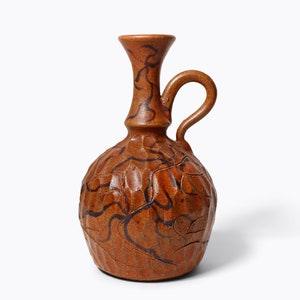 May include: A brown ceramic vase with a textured surface and a black floral design. The vase has a narrow neck and a round body with a handle.