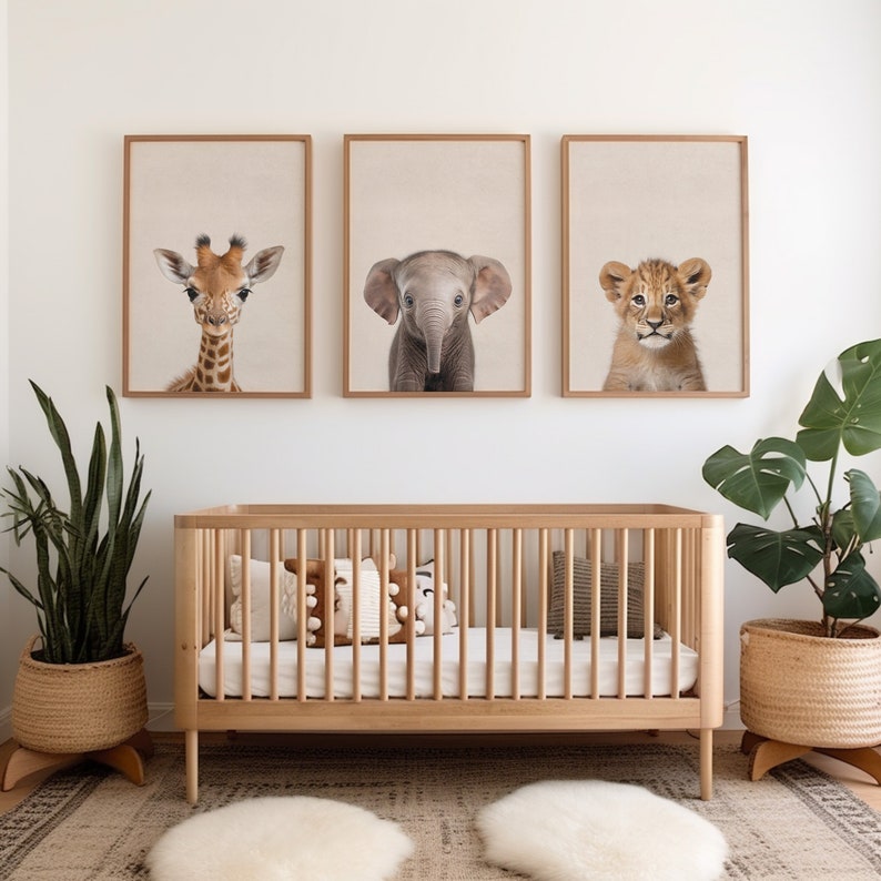 May include: Three framed animal prints of a giraffe, elephant, and lion cub, hanging above a wooden crib with a white mattress and two pillows. The crib is in a nursery with a green plant in a woven basket on the left and a monstera plant in a woven basket on the right.