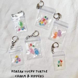 Peut inclure: Six pochettes en plastique transparent avec des porte-clés en métal argenté, contenant chacune de petites breloques en forme de tortue colorées et des perles. Le texte KOREAN LUCKY TURTLE CHARM & HIPPERS est imprimé en noir sous les pochettes.