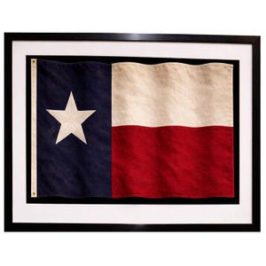 Può includere: Bandiera del Texas incorniciata, con una stella bianca su sfondo blu, una striscia bianca e una striscia rossa. La bandiera è presentata in una cornice nera con un passe-partout bianco.