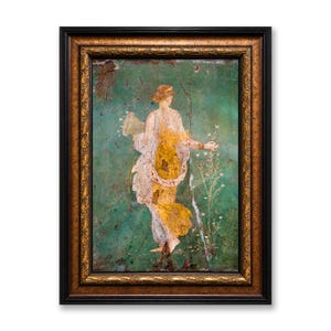 May include: A framed wall art piece featuring a weathered fresco painting of a figure in a yellow and white garment against a green background. The figure holds a cornucopia and stands near tall white flowers. The frame is ornate, dark gold and black.
