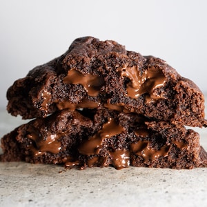 May include: Two chocolate chip cookies with melted chocolate filling. The cookies are stacked on top of each other and are slightly broken open, revealing the gooey chocolate inside.