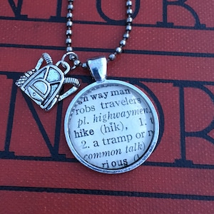 Pode incluir: Colar prateado com um pingente redondo com a definição de "hike" de um dicionário e um pequeno pingente de mochila. A corrente é de contas, ideal para caminhantes.