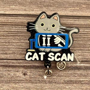 May include: A black and blue badge holder with a gray cat cartoon. The cat is inside an x-ray machine and the text "CAT SCAN" is below the image.