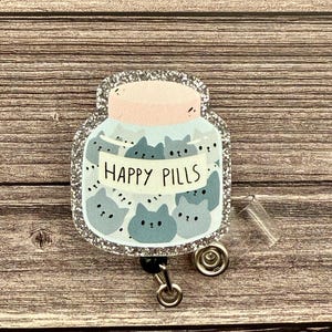 May include: A badge reel featuring a jar of "Happy Pills" with cute cartoon cat faces. The jar is light blue with a pink lid and a silver glitter border. The reel has a clear plastic retractable cord and a metal clip.