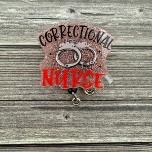 May include: A pink glitter badge holder with a black and silver retractable badge reel. The badge holder features a black and white illustration of handcuffs and the text "Correctional Nurse" in red.