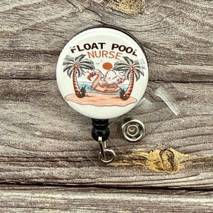 May include: White round badge holder with a black retractable cord. The badge features a cartoon flamingo on a float in a pool with palm trees and the text "Float Pool Nurse".