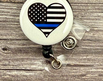 May include: A white round badge holder with a blue line heart design. The heart is split in half, with the top half being the American flag and the bottom half being a black and white striped pattern. The badge holder has a clear plastic retractable reel.