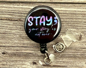 May include: A black retractable badge holder with a round, black button featuring the text "STAY; your story is not over" in a colorful, glittery font.