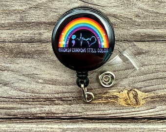 May include: A black retractable badge holder with a round button featuring a rainbow, a semicolon, a heartbeat, a heart, and the text "Broken Crayons Still Color".